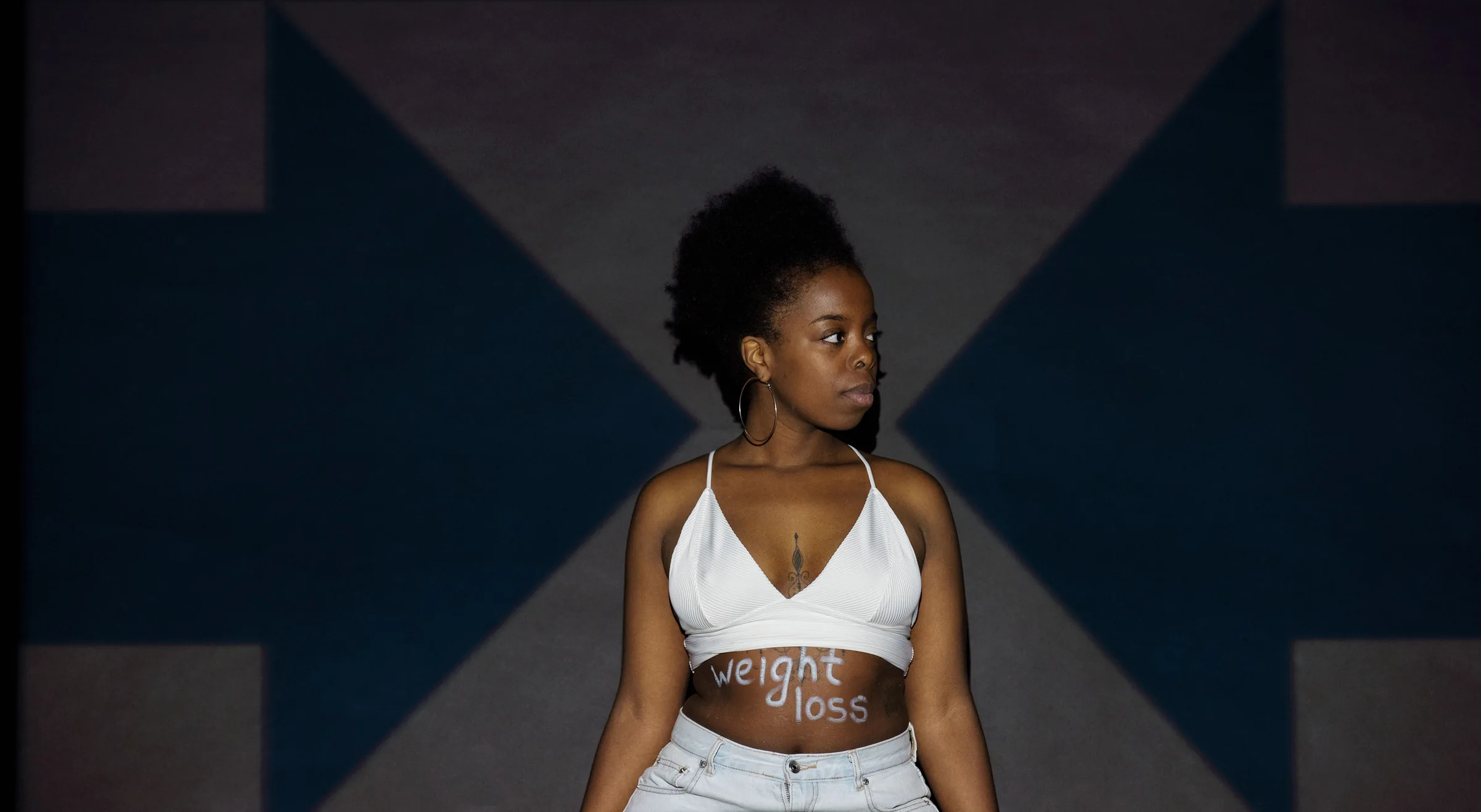 Woman in white crop top and jeans with weight loss text on her stomach against geometric blue background.